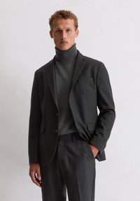 Heren donkergrijs maatpak jas met ingesneden revers, gecombineerd met een hoge hals sweater. Het materiaal heeft een subtiele textuur en patroon.