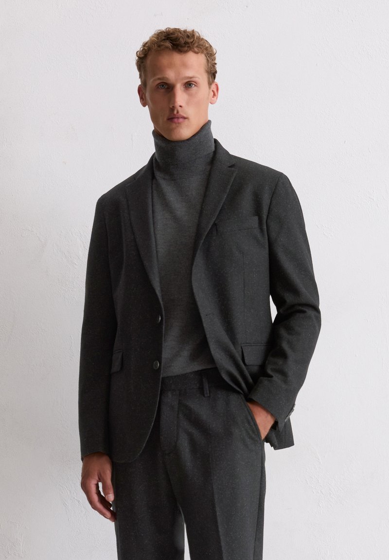 Heren donkergrijs maatpak jas met ingesneden revers, gecombineerd met een hoge hals sweater. Het materiaal heeft een subtiele textuur en patroon.