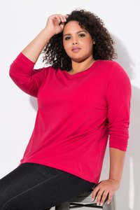 Donna con i capelli ricci che indossa una camicia rossa a maniche lunghe e pantaloni neri, seduta su uno sgabello di metallo contro uno sfondo bianco.