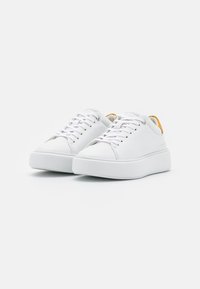 Baskets blanches avec une tige en cuir lisse, une semelle en caoutchouc texturée, des lacets plats et un accent jaune au talon. Design à bout arrondi.