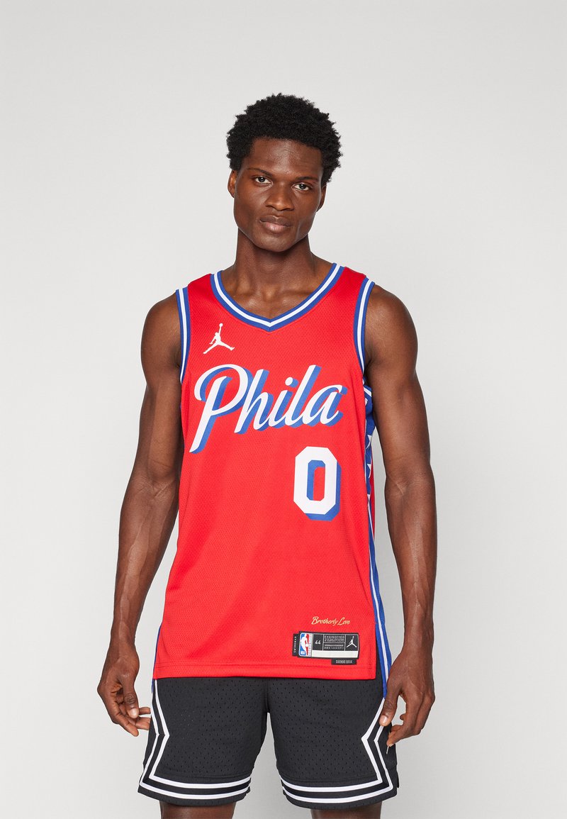 Canottiera da basket rossa con bordi blu. Presenta la scritta "Phila" in corsivo bianco e il numero "0". Tessuto a rete testurizzato e etichetta del logo NBA sull'orlo.