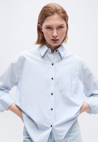 Νεαρή γυναίκα με ίσια κόκκινα μαλλιά φοράει άνετο, μακρυμάνικο πουκάμισο με κατακόρυφες λευκές και ανοιχτό μπλε ρίγες και τσέπη στο μπροστινό μέρος, με τα χέρια στη μέση.