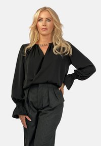 Czarna bluzka z dekoltem w kształcie litery V i zgromadzonymi mankietami, wykonana z gładkiego materiału. Połączona z spodniami w kolorze węgla z wysokim stanem w prążki. Złoty akcent w postaci łańcucha.