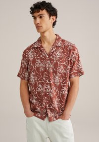 Chemise à manches courtes à motifs floraux marron avec des accents blancs, dotée d'un col et d'une coupe décontractée. Le tissu a une texture lisse.