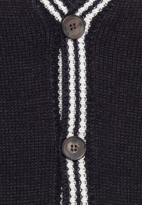 Marinblå stickad kardigan med två vita vertikala ränder, en rund brun knapp och strukturerad tyg.