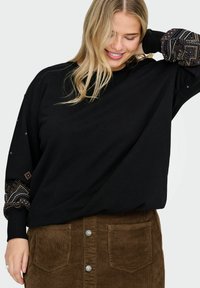 Melna sporta jaka ar rakstainām piedurknēm, kas iezīmē ģeometriskus dizainus. Saskaņota ar brūnu korduroju svārku, kura uzsvars ir uz pogu detaļām un kabatām.