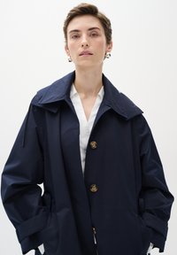 Jūras zilas ūdensnecaurlaidīgas jakas ar lielu apkakli, priekšējiem poga un sānu kabatām; iezīmē brīvu siluetu un gludu tekstūru.