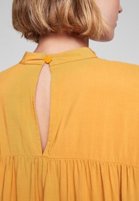 Gul bomullsklänning med ett nyckelhål i ryggen, knappstängning och draperad kjoldetalj. Mjuk textur med ett lätt utseende.