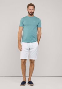 Camiseta de manga corta turquesa, pantalones cortos blancos y zapatillas sin cordones marinas. La camiseta presenta un logotipo sutil en el lado izquierdo.