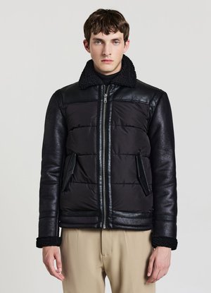 Junger Mann trägt eine schwarze Steppjacke mit Lederärmeln und Kragen, steht vor einem schlichten hellgrauen Hintergrund.