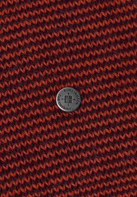 Tissu tricoté en bordeaux foncé avec des accents orange, ornée d'un bouton rond noir embossé de l'inscription « BURLINGTON THE ORIGINAL ».