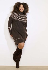 Moteris su garbanotais plaukais šypsosi, vilki rudą raštuotą mezginį suknelę ir derančius aukštus batelius iki kelių, stovi prie baltos sienos.