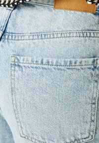 Ljust blå denimjeans med en enda bakficka, synlig sömnad och en brun läderetikett. Tyget har en något blekt textur.