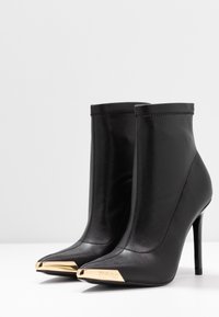 Czarne skórzane botki na wysokim obcasie sięgające kostki, z lekko spiczastymi noskami, ozdobione złotymi metalowymi czapeczkami z wygrawerowaną nazwą marki projektanta.