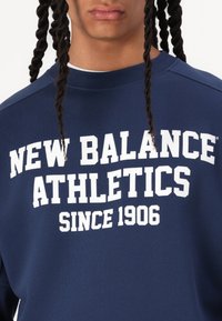 Sötétkék sweatshirt, amelyen fehér betűvel szerepel a "NEW BALANCE ATHLETICS SINCE 1906" felirat. Az anyag puha, kerek nyakkivágással és laza fazonnal rendelkezik.
