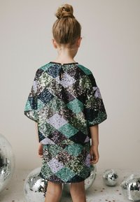 Paljettklänning i mönster av diamanter med gröna, svarta och lila nyanser; korta ärmar; nyckelhål bak; bärs av ett barn.