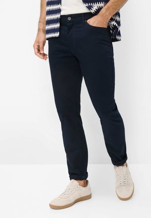Homme portant un pantalon slim bleu marine foncé, des baskets blanches et une chemise à motif bleu et blanc, les mains dans les poches.