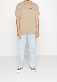 Mężczyzna w beżowej koszulce Lyle & Scott, jasnoniebieskich dżinsach i oliwkowych sneakersach stoi na tle prostej białej ściany.