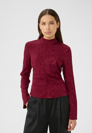 Burgundy long-sleeve blouse with textured rose pattern, mock neck, and gathered detailing at the waist, paired with black tailored trousers.