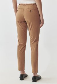 Bruine chinos met een getailleerd ontwerp, een gladde textuur en twee achterzakken met knopen. Gestoeld met zwart schoeisel.