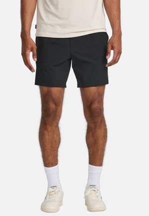 Schwarze Shorts mit einer figurbetonten Passform, aus leichtem Material; mit einem seitlichen Logo-Patch und flachem Vorderteil. Getragen mit weißen Turnschuhen und Socken.