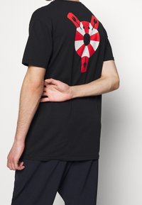 Černé bavlněné tričko s velkým červeným a bílým kruhovým grafickým motivem záchranného kruhu na zadní straně. Krátké rukávy a volný střih.