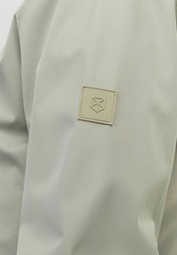 Hele roheline kangas sujuva tekstuuriga, millel on vasakul õlal ruudukujuline beež plekk ja reljeefne logo.