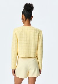 Giacca cropped in tweed giallo con una vestibilità ampia, motivo testurizzato e maniche lunghe. Abbinata a pantaloni corti giallo chiaro, che mostrano un design su misura.