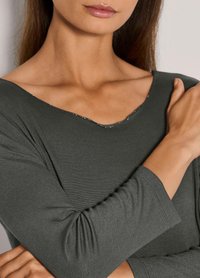 Frau trägt ein dunkelgraues Langarmoberteil mit subtiler V-Ausschnitt, mit verschränkten Armen, zeigt Hals und unteren Teil des Gesichts mit neutralen Lippen.
