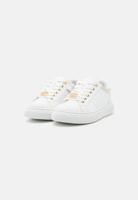 Zapatillas blancas con textura de cuero suave, con acentos de hardware dorado, puntera redonda y detalles perforados en los laterales.
