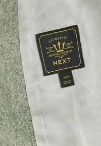 Grün und grau strukturiertes Gewebe mit einem schwarzen Etikett, das "Authentic Heritage Tailoring NEXT" liest. Größenetikett 40R, hergestellt in Vietnam.