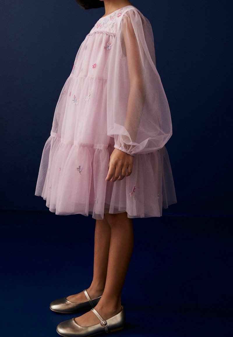 Rosa Tüllkleid mit Puffärmeln, geschichteter Gestaltung und floralem Stickerei. Kombiniert mit metallisch silbernen Ballerinas und einer runden Schnalle.