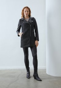 Cappotto nero con una parte anteriore in tessuto testurizzato e maniche in pelle, dotato di chiusura con zip, tasche laterali, abbinato a pantaloni e stivali in pelle nera.