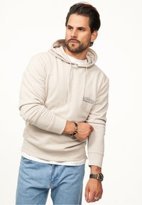 Light beige hoodie with a front pocket, drawstring hood, and embroidered logo. Paired with blue denim jeans and accessorized with bracelets.