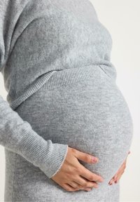 En gravid persons händer vilar på deras rundade mage, iklädd en mjuk grå tröja med ribbade ärmar. Fokuset ligger på midjan.