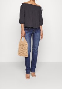 Mörk av-axlad topp med volang, jeans i denim och en beige vävd väska med trähandtag. Tydlig textur kontrasterar med den släta tyget.