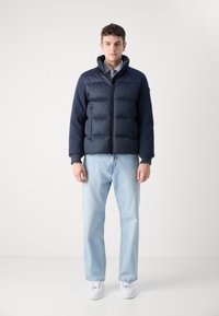 Ναυτικό μπουφάν με κουκούλα, εμπρός φερμουάρ και ραμμένη υφή. Συνδυασμένο με ανοιχτό μπλε τζιν και λευκά αθλητικά παπούτσια για μια καθημερινή εμφάνιση.