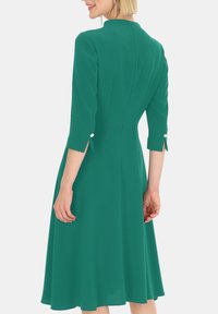 Zielona sukienka z rękawami 3/4, dopasowaną górą, rozszerzanym dołem i zamkiem błyskawicznym z tyłu. Posiada ozdobne guziki na otworach rękawów.