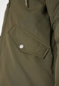 Veste verte en tissu texturé, avec une poche diagonale à fermeture à pression argentée et des accentuations de couture visibles.