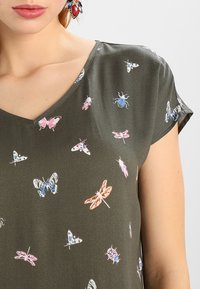 Nahaufnahme einer Frau, die eine grüne V-Ausschnitt-Bluse trägt, die mit bunten Schmetterlingen und Insekten gemustert ist, sowie mit bunten dekorativen Ohrringen.