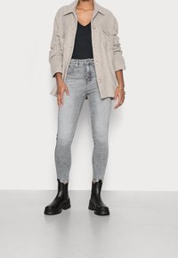 Beige knäppt jacka, svart topp, grå tajta jeans och svarta ankelskor med kraftiga sulor. Fabrikens texturer synliga i lager.