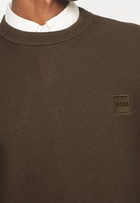 Brun, texturerad tröja med rund halsringning, som har en liten fyrkantig logotyp på bröstet i en mörkare nyans av brunt.