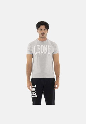 Homme debout portant un t-shirt gris clair avec le texte "LEONE 1947" et un pantalon noir avec le texte "LEONE" sur la cuisse gauche.