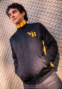 Zwarte zip-up jack met gele accenten, voorzien van een logo op de borst. De stof lijkt soepel, met zijzakken en geribde manchetten.
