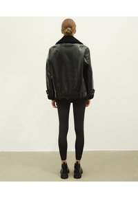Czarna kurtka z sztucznej skóry o teksturowanym wykończeniu, z kołnierzem i mankietami z baranka, noszona na czarnych legginsach i masywnych czarnych botkach.