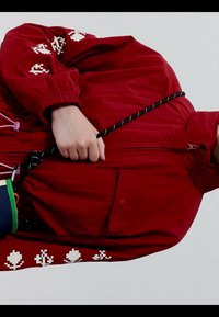 Osoba w czerwonej kurtce z białymi kwiatowymi wzorami na rękawach, trzymająca czarną linę lub sznur przeciągnięty przez klatkę piersiową.