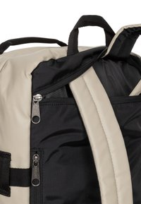 Tvåfärgad ryggsäck i cream och svart, med tygdragkedjor och funktionella handtag, en strukturerad yta och flera fack.