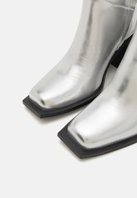 Bottines métalliques argentées avec un bout carré et un talon bloc en caoutchouc noir. Texture lisse avec une surface réfléchissante élégante.