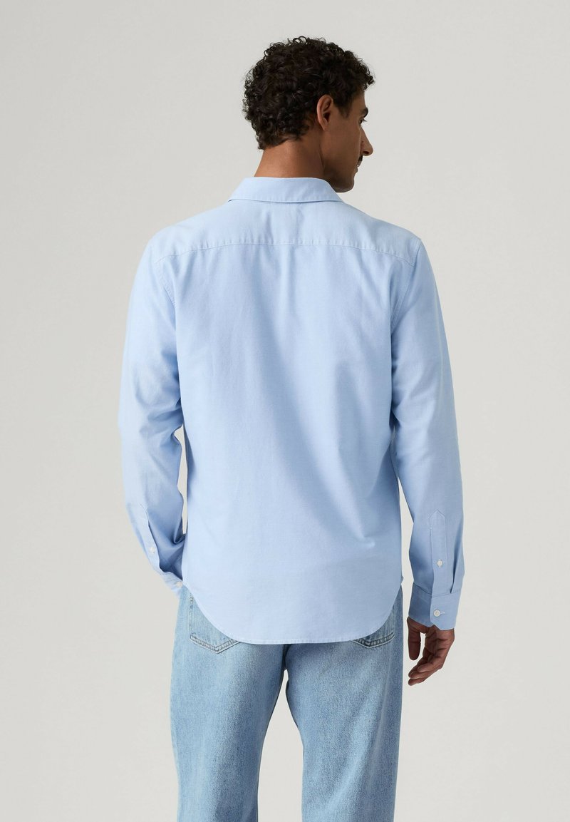 Mand iført lyseblå skjorte med knapper og lyseblå jeans set bagfra mod ensfarvet grå baggrund.