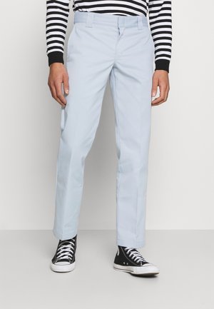 Personne portant un pantalon bleu clair à jambes droites, des baskets montantes noires et un t-shirt à manches longues rayé noir et blanc, debout contre un mur uni.